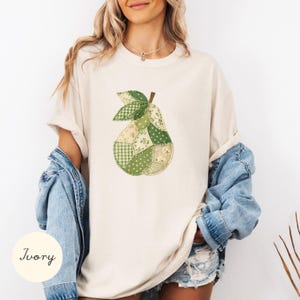 Op de afbeelding: Een ivoorkleurig t-shirt met een centrale afbeelding van een peer, opgebouwd uit diverse groene en crèmekleurige stoffen lapjes met uiteenlopende patronen zoals vichy, bloemen en stippen, gedragen over een lichtblauw spijkerjack.