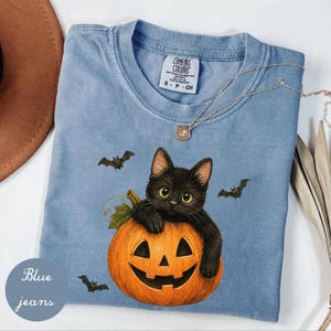 Op de afbeelding: Een blauw t-shirt met korte mouwen en een afbeelding van een zwarte kat die in een uitgesneden oranje pompoen zit. Het ontwerp bevat drie kleine zwarte vleermuizen die rond de kat vliegen. Comfort Colors label bij de hals.