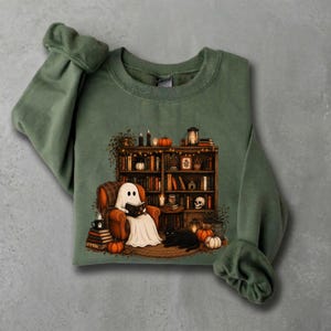 May include: An olive green crewneck sweatshirt featuring a graphic print of a white ghost sitting in an orange armchair reading a book. The scene includes a wooden bookshelf filled with books, pumpkins, a skull, a black cat, and lit candles.