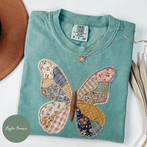Op de afbeelding: Een lichtgroen t-shirt met een grote vlinderapplicatie gemaakt van diverse stoffen met patronen, waaronder bloemen-, ruitjes- en kersenprints. Het shirt heeft een Comfort Colors-label in de nek en een dunne zilverkleurige ketting.