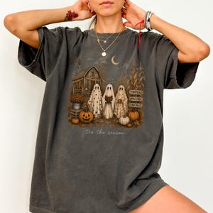 May include: A charcoal gray t-shirt featuring a graphic of three ghosts standing in front of a barn with pumpkins and corn stalks. The text reads "PUMPKINS", "APPLE CIDER", "CORN MAZE", and "tis the season" in a cursive script below the illustration.