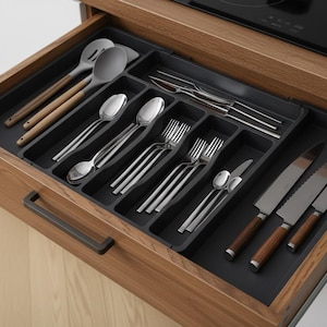 May include: A dark gray plastic kitchen drawer organizer with multiple compartments holding various stainless steel utensils, including spoons, forks, knives, and cooking tools with wooden handles, all neatly arranged inside a wooden drawer.