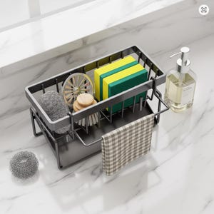 Peut inclure: Un organiseur d'évier en métal gris foncé avec un porte-éponge intégré, un distributeur de savon et une petite serviette à carreaux. L'organisateur contient des éponges, des brosses à récurer et une boule à récurer. Le distributeur de savon est transparent.