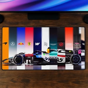 May include: A rectangular desk mat featuring a horizontal collage of Formula 1 racing car segments, each displayed against a vertical colour-blocked background representing various team liveries, logos, and sponsor branding across the full width.