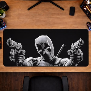 May include: Black rectangular desk mat featuring a grayscale image of Deadpool holding two pistols. The image is centered on the mat, with the character's face and weapons prominently displayed. The mat is designed for use with a computer.