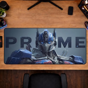 以下が含まれることがあります： Transformersのオプティマスプライムの詳細な画像が特徴の長方形マウスパッド。大きなグレーの文字で「PRIME」と書かれています。デザインは、キャラクターの青と銀色の配色を示しています。