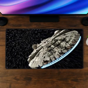 Op de afbeelding: Een rechthoekige bureaumat met een gedetailleerde afbeelding van het Millennium Falcon ruimteschip dat door de ruimte vliegt. Het ontwerp toont het grijze, cirkelvormige schip tegen een zwarte achtergrond met witte strepen die sterren voorstellen.