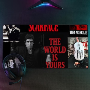Puede incluir: Una alfombrilla de escritorio rectangular con temática de la película Scarface, con un retrato en blanco y negro de Al Pacino, el texto rojo Scarface y el texto rojo The World Is Yours. Un ratón gaming blanco y auriculares negros sobre ella.