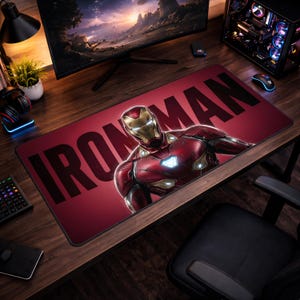 May include: A rectangular red mousepad featuring the text "IRON MAN" in large black letters. The mousepad has an image of Iron Man in his red and gold armor. The mousepad is on a wooden desk.