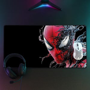 Op de afbeelding: Een rechthoekige bureaumat met een afbeelding van een rood gemaskerd personage dat gedeeltelijk bedekt is met een zwarte, stroperige substantie. Een witte, geperforeerde gamingmuis ligt op de mat en een zwarte headset met microfoon ligt ernaast.
