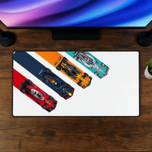May include: A rectangular desk mat featuring three Formula 1 race cars in a diagonal arrangement. The cars are positioned on colored stripes of red, dark blue with Honda branding, and orange, with a silver car on a white background.
