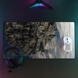 May include: A rectangular desk mat featuring a detailed graphic of the Millennium Falcon spaceship flying above a planet. A white honeycomb-patterned computer mouse and black over-ear headphones with a microphone are placed on the mat.