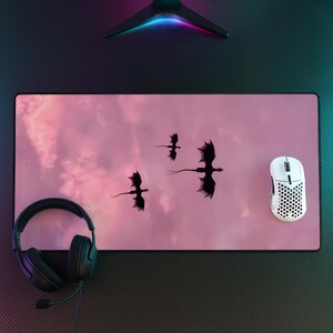 May include: A rectangular desk mat featuring a soft pink, cloud-like background with three black silhouettes of flying dragons. The mat is positioned on a dark surface with a black headset on the left and a white, perforated gaming mouse on the right.