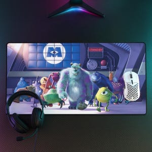 Op de afbeelding: Een rechthoekige bureaumat met een kleurrijke Monsters en Co. afbeelding met Sulley en Mike Wazowski, vergezeld door een zwarte bedrade headset en een witte geperforeerde gamingmuis. De achtergrond toont een fabrieksscène met een Stand By Scare bord.