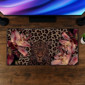May include: A rectangular desk mat featuring a central leopard head surrounded by large pink lily flowers against a tan and brown leopard print background. The mat is positioned on a dark wood surface beneath a computer monitor.
