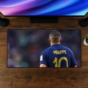 Può includere: Un tappetino da scrivania rettangolare con una stampa ad alta risoluzione di un calciatore visto di spalle. Il giocatore indossa una maglia blu scuro con il nome MBAPPE e il numero 10 stampati in lettere dorate su uno sfondo di stadio sfocato.