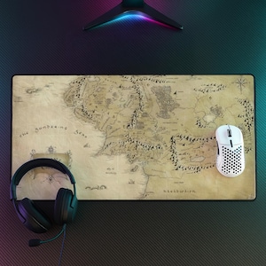 Può includere: Un tappetino da scrivania rettangolare con una mappa dettagliata in stile vintage di un mondo fantastico su sfondo beige color pergamena, illustrazioni a inchiostro nero di montagne, foreste e testo, con cuffie nere e un mouse bianco.