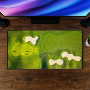 May include: A rectangular desk mat featuring an aerial view of a golf course. The design displays vibrant green grass, contoured fairways, and three distinct white sand bunkers. The mat has a black border and is positioned on a dark wood surface.