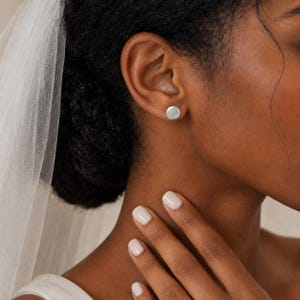 May include: A small, round, silver-toned stud earring with a smooth, polished surface. The earring is worn in the earlobe, featuring a simple, circular design that sits flush against the skin, providing a clean and understated appearance.