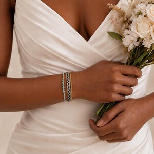 May include: A set of three bracelets worn on the wrist, including one with small light blue beads, one with a black and white chevron pattern, and one thin gold-toned chain, paired with a bouquet of white roses and small filler flowers.