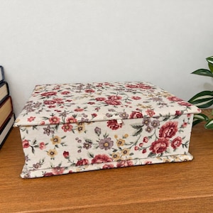 May include: A rectangular fabric-covered storage box featuring a cream background with a floral pattern of red, pink, yellow, and purple flowers and green leaves. The box has a flat lid and a small fabric tab for opening the lid.