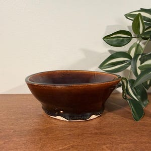 May include: A round ceramic bowl featuring a deep brown, glossy glaze that transitions to a darker, almost black tone near the base. The bowl has a wide, open rim and a smooth, reflective surface, showcasing a simple, functional design.
