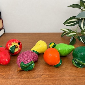 May include: A collection of six ceramic fruit figurines including a red strawberry, a red apple, a purple raspberry, a yellow pear, an orange, and a green lime. Each piece features a glossy finish and painted green leaf details.