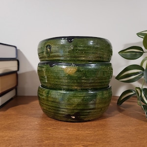 May include: A stack of three round ceramic bowls with a glossy, mottled forest green glaze. The bowls feature a horizontal ribbed texture along the exterior sides, with subtle variations in color including dark green and hints of yellow-brown.