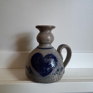 May include: A speckled gray ceramic candle holder with a bulbous base and a single handle. The piece features a prominent dark blue heart design on the front, a blue horizontal line above the heart, and a flared top rim for holding a candle.