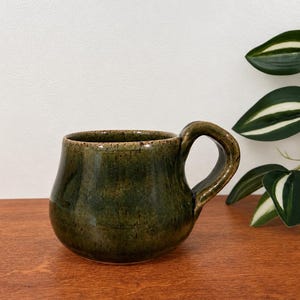 May include: A handmade ceramic mug with a rounded, bulbous body and a thick, curved handle. The mug features a mottled, dark olive green glaze with subtle brown speckles throughout the surface, creating a textured and earthy appearance.