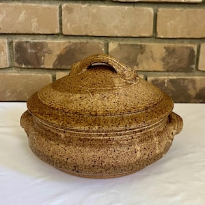 May include: A round, lidded ceramic casserole dish featuring a speckled tan and brown glaze. The pot has two small side handles and a matching lid with a central arched handle, showcasing a textured, earthy finish throughout the entire piece.
