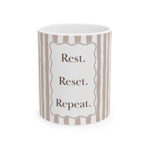 Könnte beinhalten: Ein weißer Keramikbecher mit vertikalen hellbraunen Streifen und einem mittigen, wellenförmig umrandeten rechteckigen Rahmen. Im Inneren des Rahmens steht der dunkelbraune Text: Rest. Reset. Repeat. Das Design umschließt das zylindrische Gefäß.