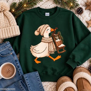 Könnte beinhalten: Ein dunkelgrüner Rundhals-Pullover mit einer grafischen Illustration einer weißen Gans, die eine beige Strickmütze und einen Schal trägt und einen kleinen, mit Tannenzweigen geschmückten Holzschlitten zieht. Die Gans hat orange Füße und einen Schnabel.