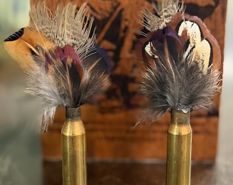 Peut inclure: Deux objets décoratifs composés de douilles de balles en laiton servant de base à des arrangements verticaux de plumes naturelles. Les plumes présentent un mélange de tons marron, beige, noir et violet irisé, fixées au sommet de chaque douille.