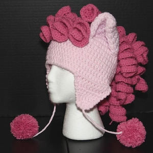May include: A light pink crocheted hat featuring ear flaps and two long braided ties ending in dark pink pom-poms. The hat is adorned with dark pink crocheted curls on the top and back, resembling a mane, and includes two pointed ears.