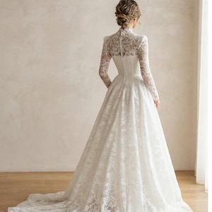 May include: A floor-length white lace gown featuring a high neckline, long sleeves, and a fitted bodice with a vertical row of small buttons running down the center back. The skirt is full and flares out into a train with intricate floral lace patterns.