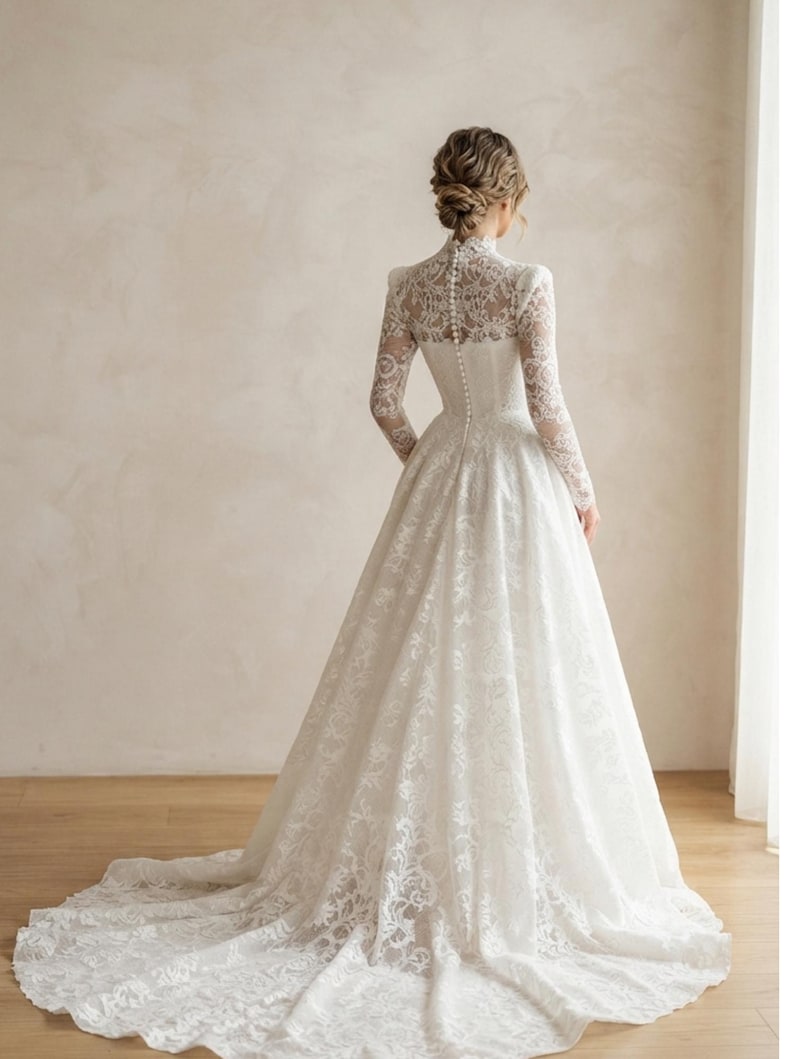 May include: A floor-length white lace gown featuring a high neckline, long sleeves, and a fitted bodice with a vertical row of small buttons running down the center back. The skirt is full and flares out into a train with intricate floral lace patterns.