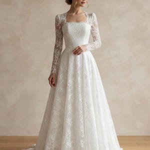 May include: A floor-length white lace gown featuring long sleeves, a square neckline, and a fitted bodice that transitions into a full, flowing skirt. The intricate floral lace pattern covers the entire garment, extending to the hemline.