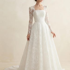 May include: A floor-length white wedding dress featuring long lace sleeves, a square neckline, and a fitted bodice that transitions into a full, flowing skirt with a train. The entire garment is covered in a delicate floral lace overlay pattern.