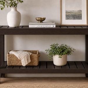 May include: A dark brown wooden console table featuring a slatted top and a lower shelf. The table has four sturdy legs and a rectangular frame. It is decorated with a white vase, a book, a small bowl, a woven basket, and a potted green plant.