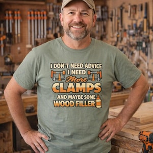 Könnte beinhalten: Ein salbeigrünes Kurzarm-T-Shirt mit dem Text I DON'T NEED ADVICE I NEED MORE CLAMPS AND MAYBE SOME WOOD FILLER in orangefarbener und weißer Schrift, ergänzt durch grafische Darstellungen von zwei orangefarbenen Schraubzwingen und einer Flasche Holzfüller.