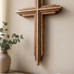 Könnte beinhalten: Ein Wandkreuz aus geschichteten Holzsegmenten in kontrastierenden hellen und dunklen Brauntönen. Das Design besteht aus einem zentralen vertikalen Balken, der von einem horizontalen Riegel gekreuzt wird und eine mehrdimensionale, gestufte Struktur bildet.