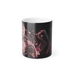 May include: A black ceramic mug featuring a monochromatic pink-toned photographic print of a person holding a microphone. The design includes geometric shapes and lines in the background, with a white interior and a smooth, glossy finish.