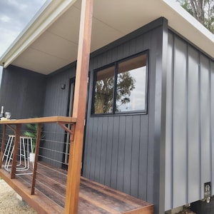May include: A small, modern cabin with dark gray vertical siding and a flat roof. It features a wooden deck with a matching railing, a sliding glass window, and a small porch area. The structure is elevated off the ground on a gravel surface.