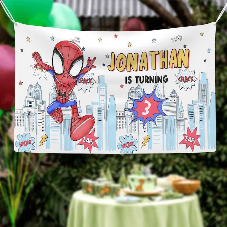 Puede incluir: Una pancarta de fiesta rectangular blanca con un personaje de Spider-Man de dibujos animados, una ilustraci&oacute;n de un horizonte urbano y el texto JONATHAN IS TURNING 3. El dise&ntilde;o incluye burbujas de c&oacute;mic con palabras como POW, CRACK y ZAP.