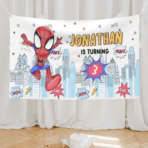 Puede incluir: Una pancarta rectangular de tela con un superh&eacute;roe de dibujos animados con traje rojo y azul saltando sobre un horizonte urbano azul claro. El texto dice JONATHAN IS TURNING 3, rodeado de burbujas de c&oacute;mic con palabras como POW, CRACK y ZAP.