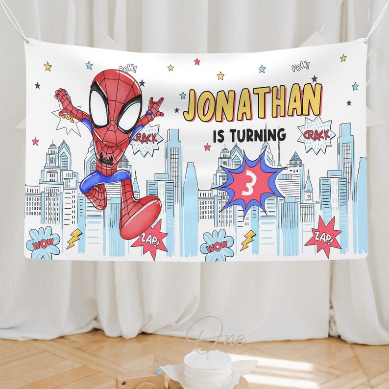 Puede incluir: Una pancarta rectangular de tela con un superh&eacute;roe de dibujos animados con traje rojo y azul saltando sobre un horizonte urbano azul claro. El texto dice JONATHAN IS TURNING 3, rodeado de burbujas de c&oacute;mic con palabras como POW, CRACK y ZAP.