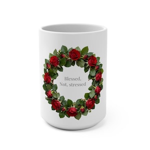 May include: A white ceramic mug featuring a circular wreath of vibrant red roses and green leaves. Centered within the floral wreath is the text "Blessed, Not, stressed" printed in a simple, dark grey, sans-serif font on the smooth surface.