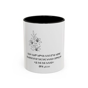 Può includere: Una tazza in ceramica bianca con interno e bordo neri. L'esterno presenta un'illustrazione a linee nere di tre gigli sopra il testo in amarico "ዕረፉ፥ እኔም አምላክ እንደ ሆንኩ እወቁ፤ በአሕዛብ ዘንድ ከፍ ከፍ እላለሁ፥ በምድርም ላይ ከፍ ከፍ እላለሁ። መዝ 46:10".