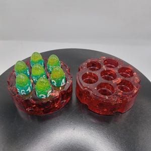 May include: A red resin revolver cylinder dice holder with gold flakes, containing six green and yellow bullet-shaped dice with white numbers. Beside it sits a matching empty red resin revolver cylinder dice holder with seven circular slots.